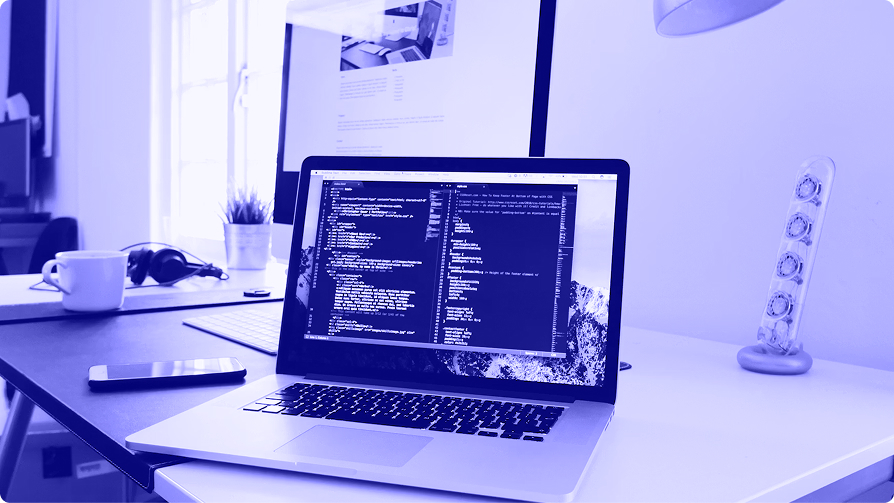 Laptop on a desk showing a split-screen code editor, with an external monitor, coffee cup, and headphones nearby.