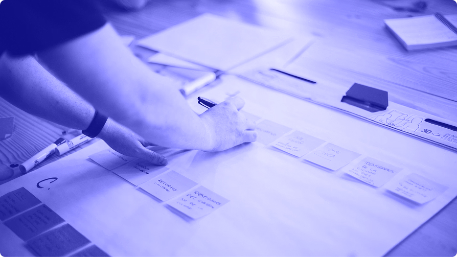 Hand holding a marker over a large sheet of paper laid out with sticky notes and hand-drawn diagrams in a planning session.