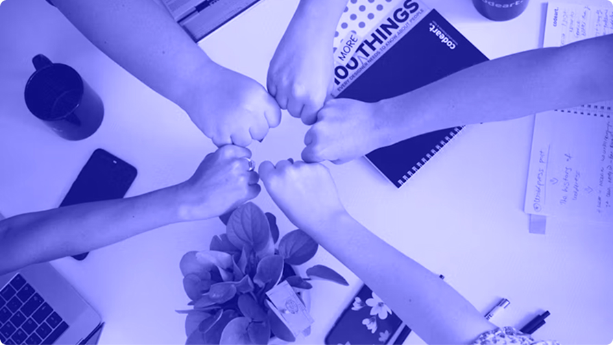 Overhead view of six people meeting their fists together in a circle above a desk of notebooks, laptops, and a plant.