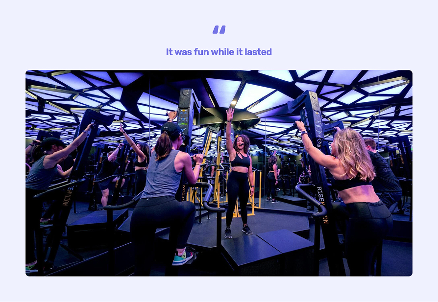 Fitness instructor in black workout clothes leads a class on step machines with mirrored walls reflecting participants raising their arms.